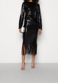 Černé šaty se flitry ve dvou kusech, které mají dlouhé rukávy, krátký top a sukni s vertikálními třásněmi na lemu. Stříbrná kabelka a sandály.
