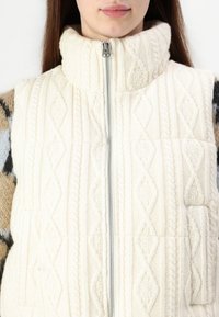 Πλεκτό κρεμ ντεσέν με ψηλό γιακά, με διαμαντένιο μοτίβο και μπροστινό φερμουάρ. Υφασμάτινη υφή με περίπλοκο σχέδιο.