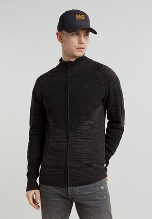 Schwarzer Zip-Up-Pullover mit strukturiertem Muster und hohem Kragen, kombiniert mit einer schwarzen Mütze mit gelbem Logo. Vor einem neutralen Hintergrund präsentiert.