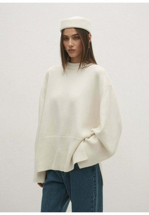 Kobieta w dużym kremowym swetrze i dopasowanym berecie, pozująca na jednolitym jasnym tle z jedną ręką w kieszeni i neutralnym wyrazem twarzy.