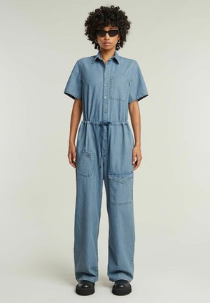 Combinaison en denim à manches courtes, avec un boutonnage à l'avant, taille cintrée, deux grandes poches avant, couleur bleu clair et coupe décontractée. Chaussures à plateforme noires.