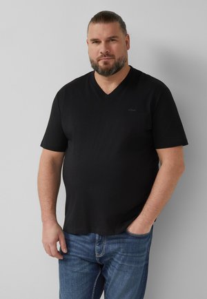 Brodaty mężczyzna z związanymi włosami, ubrany w czarną koszulkę z dekoltem w serek i niebieskie dżinsy, stojący na jednolitym jasnoszarym tle.