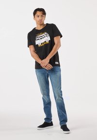 Svart T-shirt med en vit bussgrafik och gul text, kombinerad med blå jeans och svarta sneakers. Enkel design, avslappnad passform.