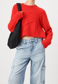 Suéter corto rojo con ribetes en el dobladillo y las mangas, que presenta un gran diseño de lazo. Combinado con jeans cargo sueltos de color azul claro y un bolso negro.