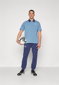 Zilā polo krekls ar tumši zilu apkakli, savienots ar tumši ziliem sporta biksēm. Modelis tur melnbaltu futbola bumbu. Izskatu papildina sporta apavi.