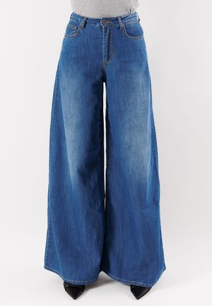 Donna con jeans di denim blu a vita alta e gamba larga e scarpe nere con punta a punta, in piedi contro uno sfondo bianco semplice.