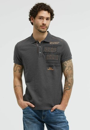 Man with curly hair wearing a dark gray polo shirt with text and tattoos on both arms, posing with hands in blue jeans pockets.