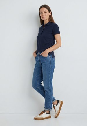 Kvinde stående med hænderne i lommerne iført navy polo shirt, blå jeans og hvide sneakers med brune accenter mod en ensartet lys baggrund.