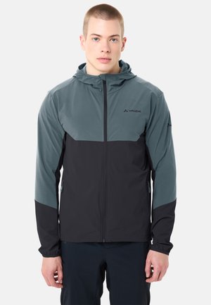 Jeune homme portant une veste à capuche Vaude zippée intégralement, bicolore bleu et noir, debout devant un fond clair uni.