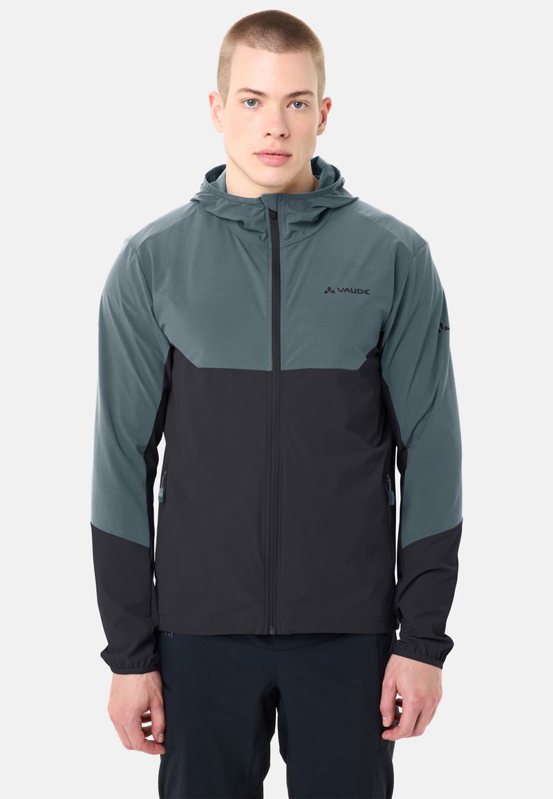 Giovane uomo che indossa una giacca con cappuccio Vaude a zip integrale bicolore blu e nera, in piedi contro uno sfondo chiaro e semplice.