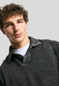 Giovane uomo con capelli ricci e una leggera peluria sul viso, che indossa una camicia col colletto grigio scuro su una T-shirt bianca, su uno sfondo neutro.