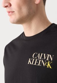 Sort bomulds T-shirt med rund hals, korte ærmer og et iøjnefaldende trykt logo i beige og gul placeret på brystet.