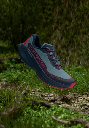 Scarpa da trail running blu e nera con accenti rossi e logo "LA SPORTIVA", sospesa sopra un terreno forestale con erba e rocce.