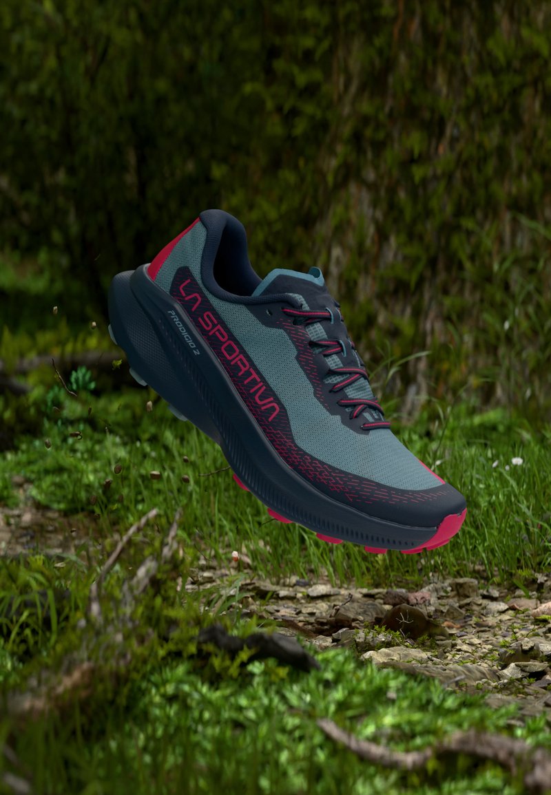 Chaussure de trail running bleue et noire avec des accents rouges et le logo "LA SPORTIVA", suspendue au-dessus d'un sol forestier avec de l'herbe et des rochers.