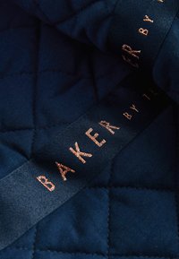 Navy gesteppter Stoff mit Rautenmuster und einem Satinband, das "BAKER by T." in Roségold geprägt zeigt. Glatte Textur und subtiler Glanz.
