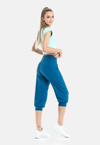 Pantalones capri en color azul verdoso con cintura alta y puños fruncidos. Combinados con un top corto verde claro y zapatillas deportivas blancas. Textura de tela suave.