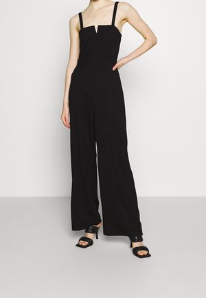 Vrouw die een mouwloze zwarte jumpsuit met wijde pijpen en een vierkante halslijn draagt, gecombineerd met zwarte sandalen met hakken, staand met één hand op haar heup.