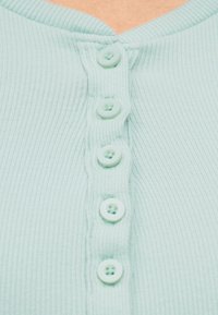 Κοντινό πλάνο ενός ανοιχτοπράσινου ριμπ πλεκτού τοπ με πέντε μικρά κουμπιά ίδιου χρώματος κατά μήκος του μπροστινού ντεκολτέ.