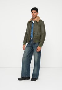Uomo in piedi che indossa una giacca bomber verde con colletto in shearling, camicia di jeans blu, jeans a gamba larga e stivali neri su sfondo bianco.
