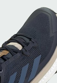 Marineblaue Sportschuhe mit strukturiertem Strick-Obermaterial, grauen Akzenten, traditionellen Schnürsenkeln und einer gepolsterten Sohle mit drei gestreifen Designelementen.