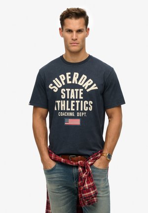 Marineblaues Baumwoll-T-Shirt mit weißem "SUPERDRY STATE ATHLETICS"-Druck und kleiner amerikanischer Flagge. Getragen mit hellblauen Jeans und einem rot karierten Hemd, das um die Taille gebunden ist.