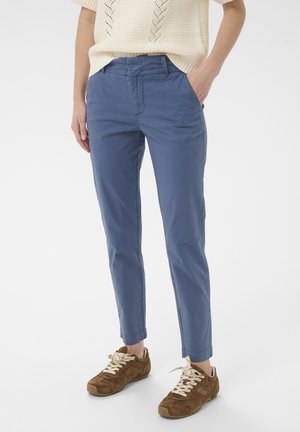 Personne portant un pantalon bleu slim, un haut en maille beige et des baskets en daim marron, debout avec une main dans la poche, sur un fond blanc.