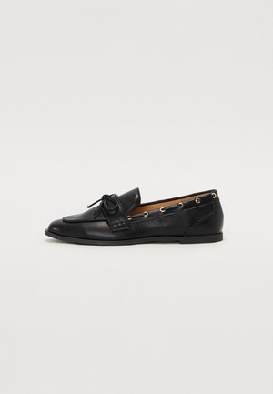 Mocassins de couro preto com um laço atado, apresentando detalhes de ilhós dourados nas laterais e uma sola de borracha plana. Textura suave, design clássico.