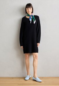 Black long-sleeve dress with a rounded neckline, paired with a multicolored silk scarf tied at the neck and light blue loafers.