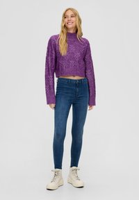 Fialový pletený zkrácený svetr s texturovaným vzorem, kombinovaný s modrými skinny džíny a krémovými vysokými teniskami. Bílý pozadí.