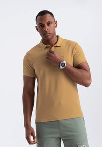 Khaki-paitainen polo-paita, joka on valmistettu puuvillasta ja jossa on kaulus sekä lyhyet hihat. Siinä on hillitty emblem rinnan kohdalla, ja se on yhdistetty cargo-shortsien kanssa.