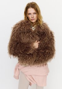 Ung kvinde med løst blondt hår iført en voluminøs brun, shaggy pelsfrakke og en lyserød striktrøje bundet om livet.