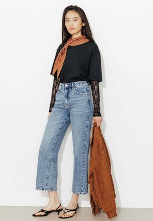 Femme portant une chemise noire avec des manches en dentelle, un jean taille haute bleu, une écharpe orange, des sandales noires à talons, tenant une veste marron sur le côté.