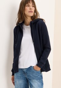 Veste à zip bleu marine avec capuche, dotée d'une texture douce, de poches latérales et de poignets côtelés, portée sur un t-shirt blanc et un jean en denim.