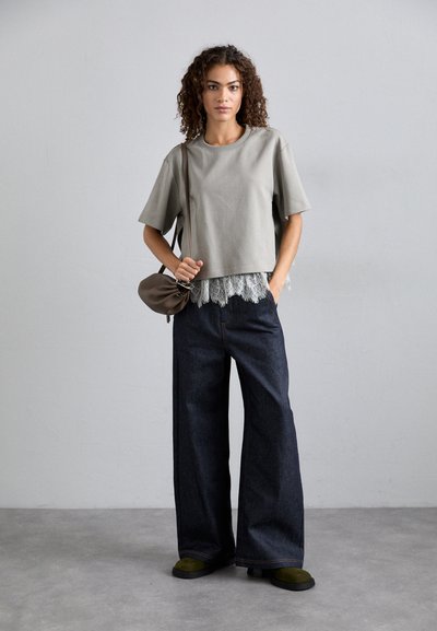 Γκρι κοντομάνικο μπλουζάκι με δαντελωτό εσωτερικό, συνδυασμένο με φαρδιές τζιν παντελόνες σε σκούρο χρώμα. Το μοντέλο κρατά μια καφέ τσάντα και φοράει παπούτσια με πράσινες λεπτομέρειες.