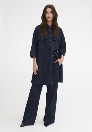 Abito camicia in denim scuro con bottoni sul davanti, due tasche sul petto e maniche corte, abbinato a pantaloni larghi in denim scuro e scarpe nere a punta.
