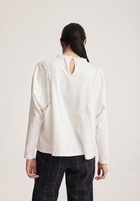 Camisa blanca de manga larga con detalles de volantes a lo largo de los hombros y una abertura en forma de ojo de cerradura en la espalda. Combinada con pantalones oscuros a cuadros.