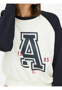 Sudadera blanca y azul marino con una gran letra "A" en negro, con "1995" en rojo. Mangas largas y sueltas y textura suave.