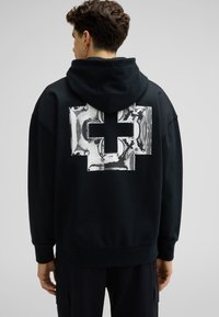 Sweat à capuche noir avec un grand design de croix métallique brillante au dos. Fabriqué en tissu doux avec une capuche à cordon et des poignets côtelés.