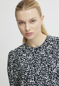 Sort-hvid mønstrede bluse med høj halsudskæring, plisseret design og knapper ned foran. Stof ser glat og let ud.