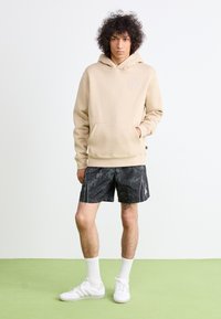 Felpa beige con cappuccio e tasca a marsupio, dettaglio del logo grigio, abbinata a pantaloni corti neri a motivo e sneakers bianche. Sfondo verde.