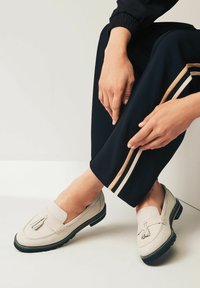 Beige nahka loaferit, joissa on tupsumerkinnät ja mustat kumipohjat, yhdistettyinä tummansinihin housuihin, joissa on kerma- ja ruskearaidat.