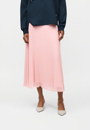 Personne portant un haut à manches longues bleu marine, une jupe midi rose clair et des talons pointus beige, debout devant un fond blanc uni.