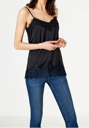 Czarna satynowa bluzka na ramiączkach z koronkowymi wykończeniami przy dekolcie i dolnej krawędzi, z regulowanymi ramiączkami i falbaniastymi brzegami. Połączona z niebieskimi dżinsami.