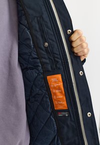 Mörkblå vadderad jacka insida med silverfärgad dragkedja, orange etikett och vindtåligt tyg. Har hållbar sömnad och isolering.