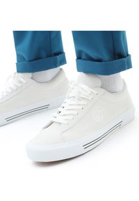 Hvite semskede sneakers med hvite snørebånd, hvit gummisåle og sidestriper. Kombinert med rullete teal-bukser og lysegrå sokker.