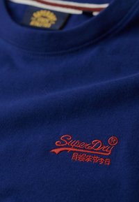 Camiseta de algodón azul marino con un logotipo "SuperDry" bordado en rojo. Cuello redondo clásico y textura suave y lisa.