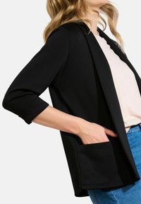 Černý blyzer vyrobený z měkké látky, s upraveným střihem, tříčtvrtečními rukávy, otevřeným předním designem a detaily předních kapes.