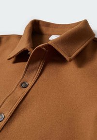 Chemise en laine marron avec col plié et boutons visibles sur la patte de boutonnage avant, présentée en gros plan sur une surface plane.