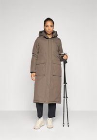 Donna con lungo cappotto marrone con cappuccio che tiene bastoni da trekking neri, in piedi contro uno sfondo grigio chiaro semplice.