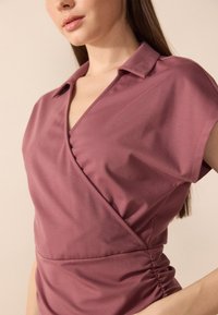 Kortermet, omslagsbluse i mauve stoff, med krage, rynket midje og jevn tekstur. Ideell for variert styling.